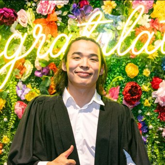 Student Erdene-Od from Mongolia smiles towards the camera. He wears a white business shirt and a black graduate cloak. He is giving a thumbs up with his right hand. Behind him is a wall covered with different coloured flowers and green leaves. Across the wall is a neon yellow light that reads Congratulations.