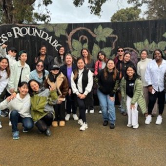 Photo displays a group of 18 international students, of different nationalities, accompanied by two adults from South Regional TAFE. Behind the group is a black wooden fence with green vines drawn artistically across the fence. In white font, placed within the vine, reads "Swings & Roundabouts." In the far distance are big trees.