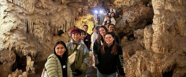 A large group of international students, with 13 in focus, stands in a line on a bridge extending from the cave's entrance. They are all smiling and looking at the camera, with the line of students continuing out of view towards the back.